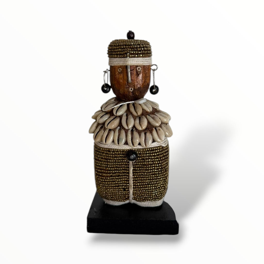 A traditional African Namji doll, made of wood and adorned with beads, cowrie shells, and metal embellishments, displayed on a white background.