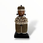 A traditional African Namji doll, made of wood and adorned with beads, cowrie shells, and metal embellishments, displayed on a white background.