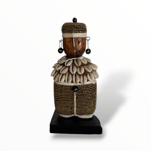 A traditional African Namji doll, made of wood and adorned with beads, cowrie shells, and metal embellishments, displayed on a white background.