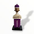 A handcrafted Namji doll made of wood, adorned with beads, cowrie shells, leather, and metal, standing on a black base.