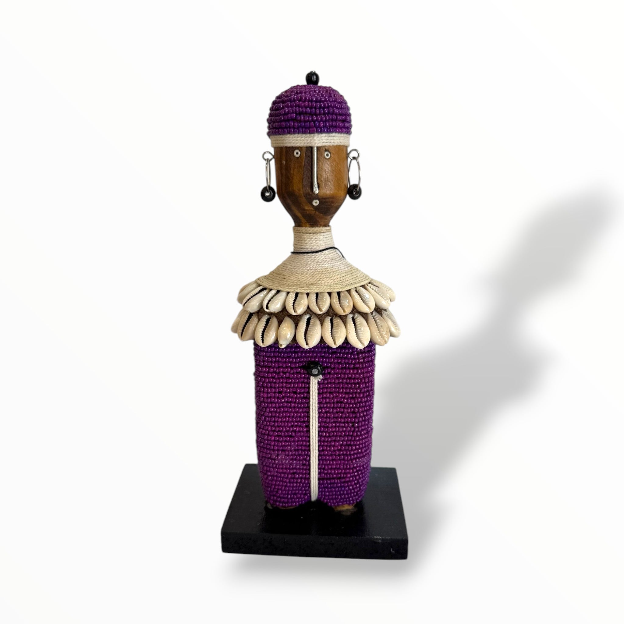 A handcrafted Namji doll made of wood, adorned with beads, cowrie shells, leather, and metal, standing on a black base.