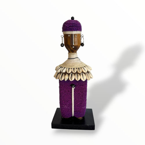 A handcrafted Namji doll made of wood, adorned with beads, cowrie shells, leather, and metal, standing on a black base.