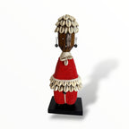 A traditional African Red Namji doll, made of wood and adorned with beads, cowrie shells, and metal embellishments, displayed on a white background.